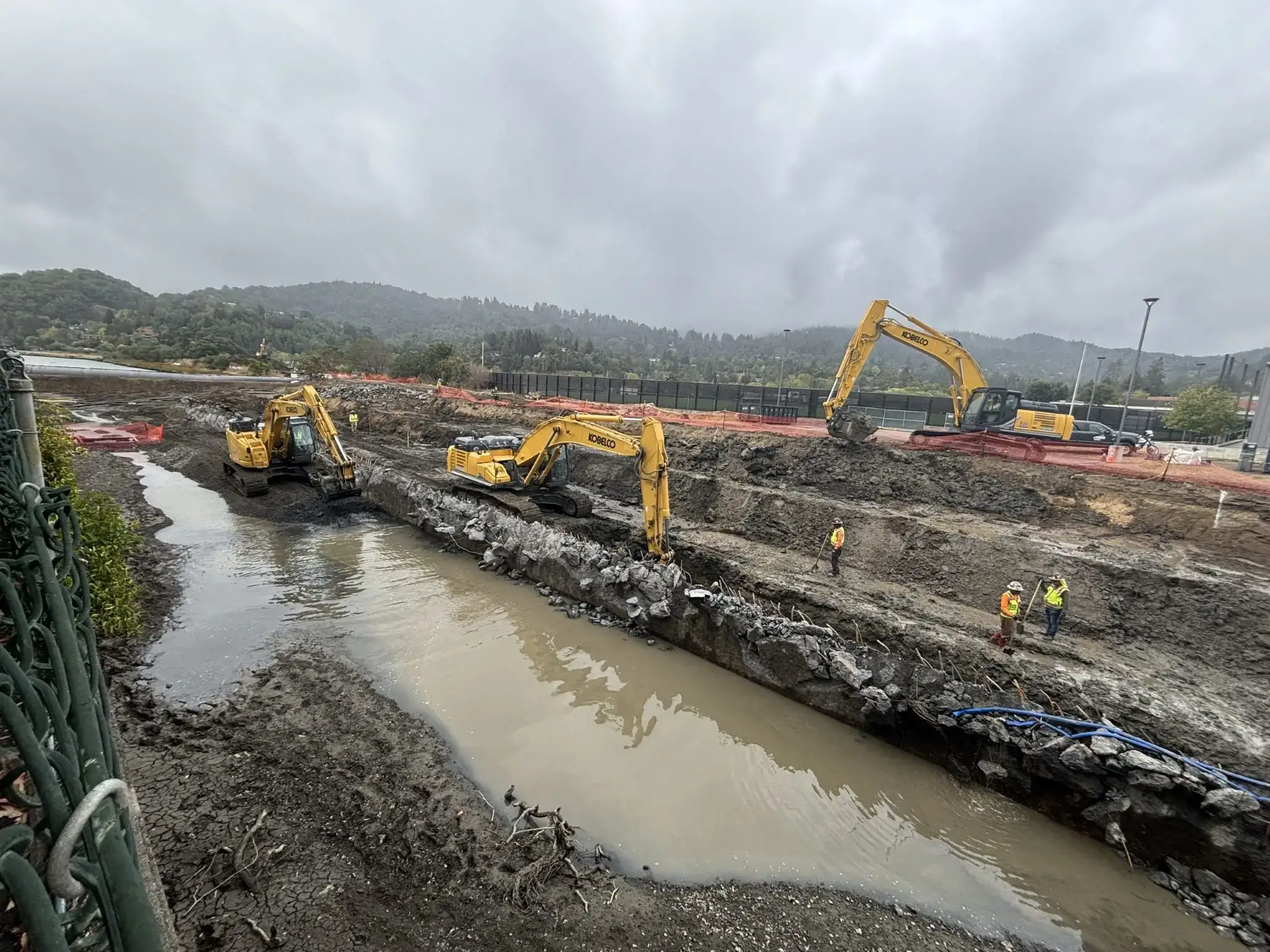 Construction vehicles working on a muddy site near a water channel.